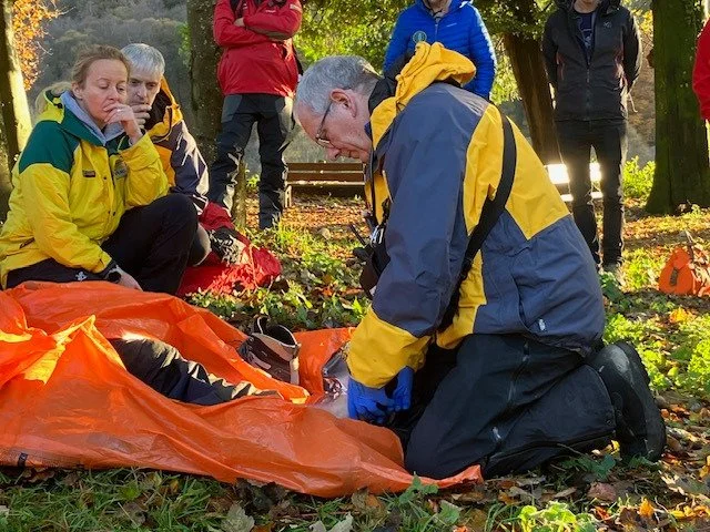 A group of outdoor enthusiasts, including a man in a yellow and gray jacket, kneels on the ground and appears to be attending to a person inside an orange rescue blanket. Others are standing or sitting nearby, observing, in a forest setting.