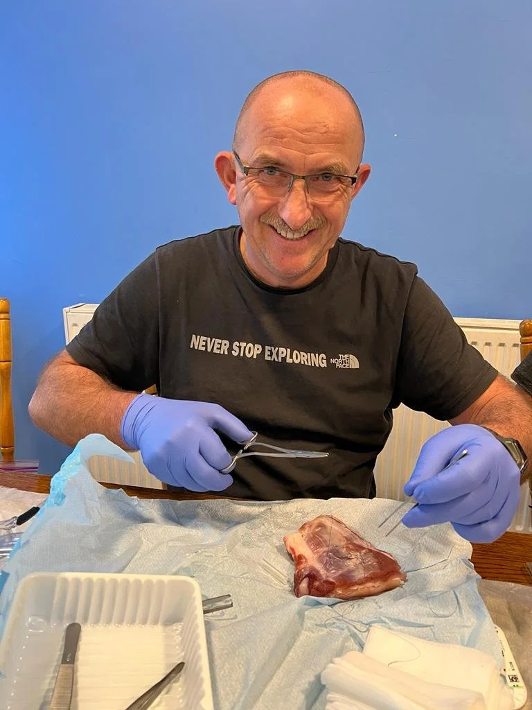 A man smiling at a table with a piece of meat, wearing a black T-shirt, glasses, and purple gloves, holding surgical tools.