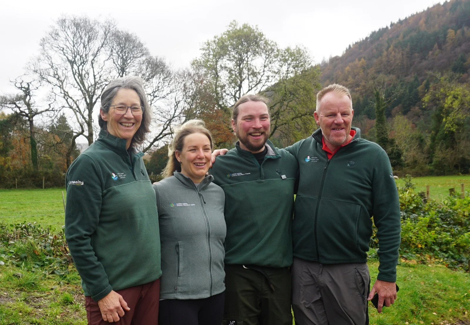 Four people standing outdoors in a grassy field with trees and hills in the background, smiling and posing for a photo, wearing outdoor jackets.