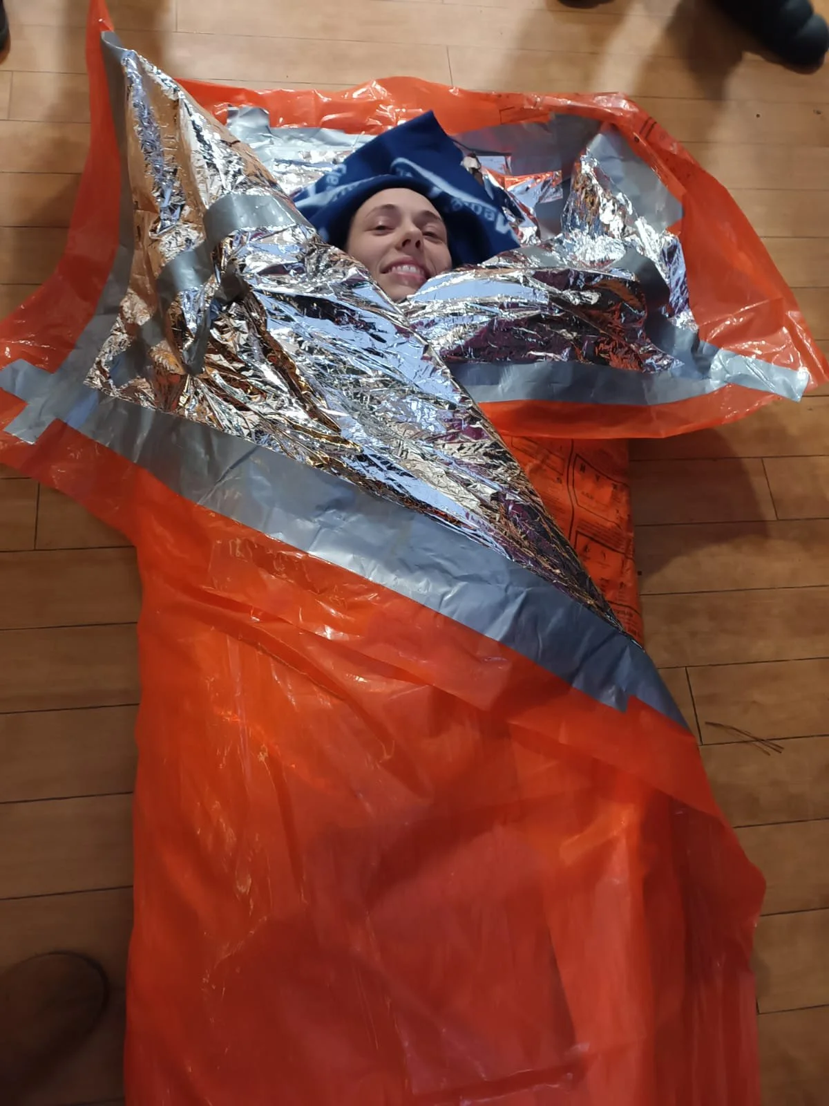 Person smiling, lying inside a space emergency blanket on a wooden floor, wrapped in an orange survival sleeping bag with reflective silver lining.