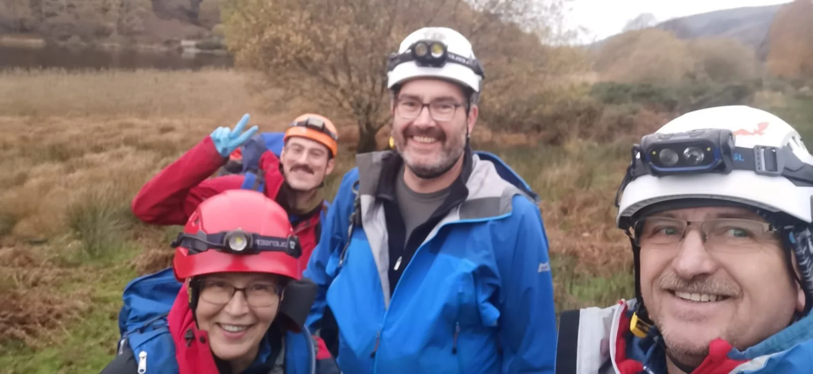 Group of four people dressed for outdoor adventure, wearing helmets with headlamps, smiling for a selfie in a grassy outdoors area with trees and hills in the background.