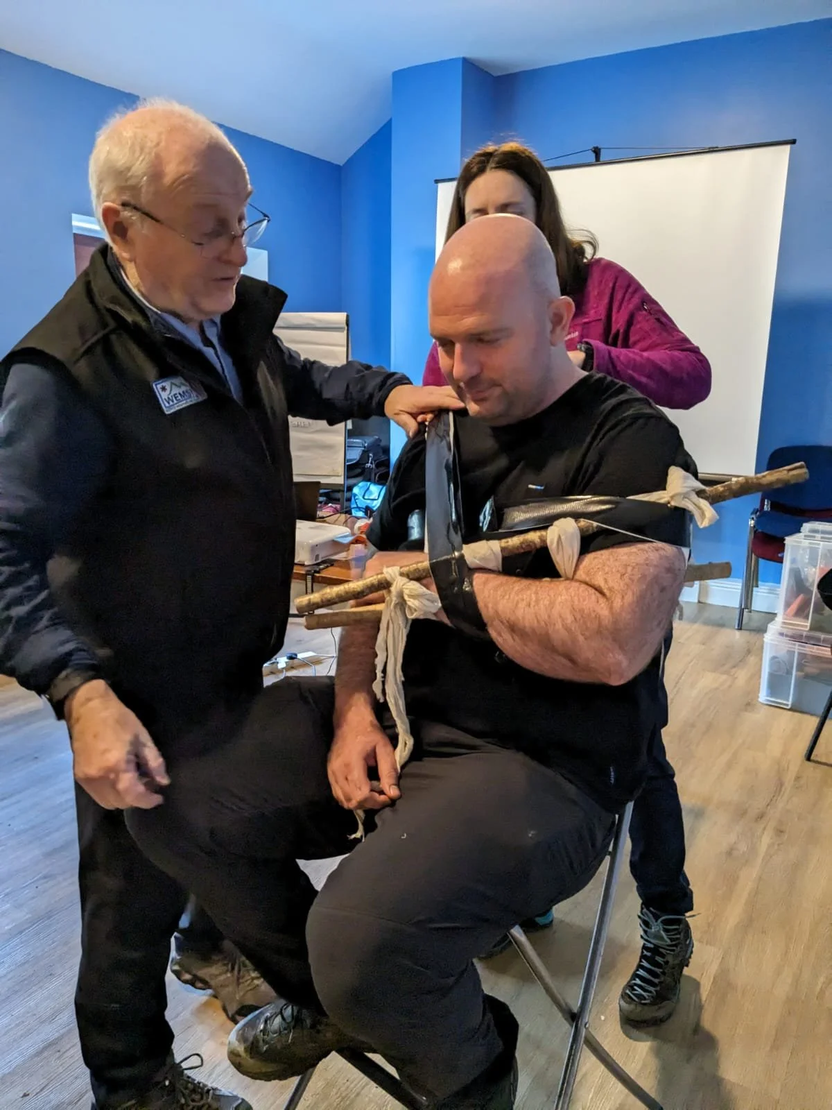 A man with a bald head seated in a chair wearing a makeshift bow and quiver, with an older woman and a younger woman assisting him in a room with blue walls. The man appears to be in a training or demonstration setting.