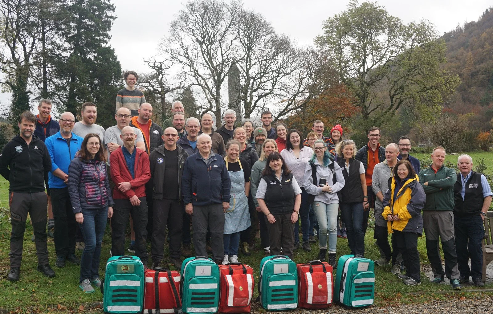 A large group of people, including men and women of various ages, standing outdoors on grass with trees and hills in the background. Some individuals are wearing outdoor or hiking clothing, and there are red and teal backpacks with labels in front of