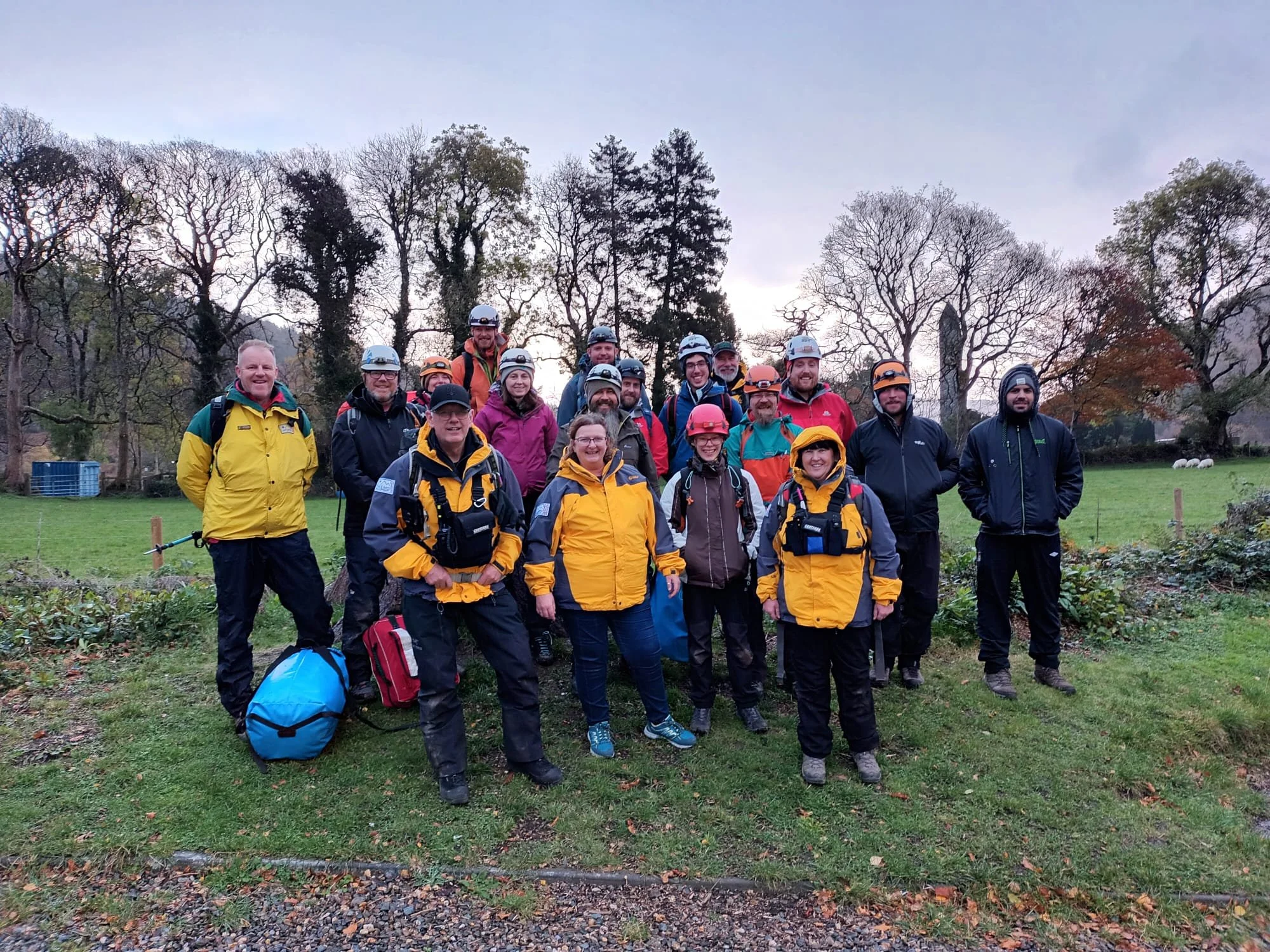 A group of people dressed in outdoor gear, wearing helmets, outdoors in a park with trees and a field in the background.