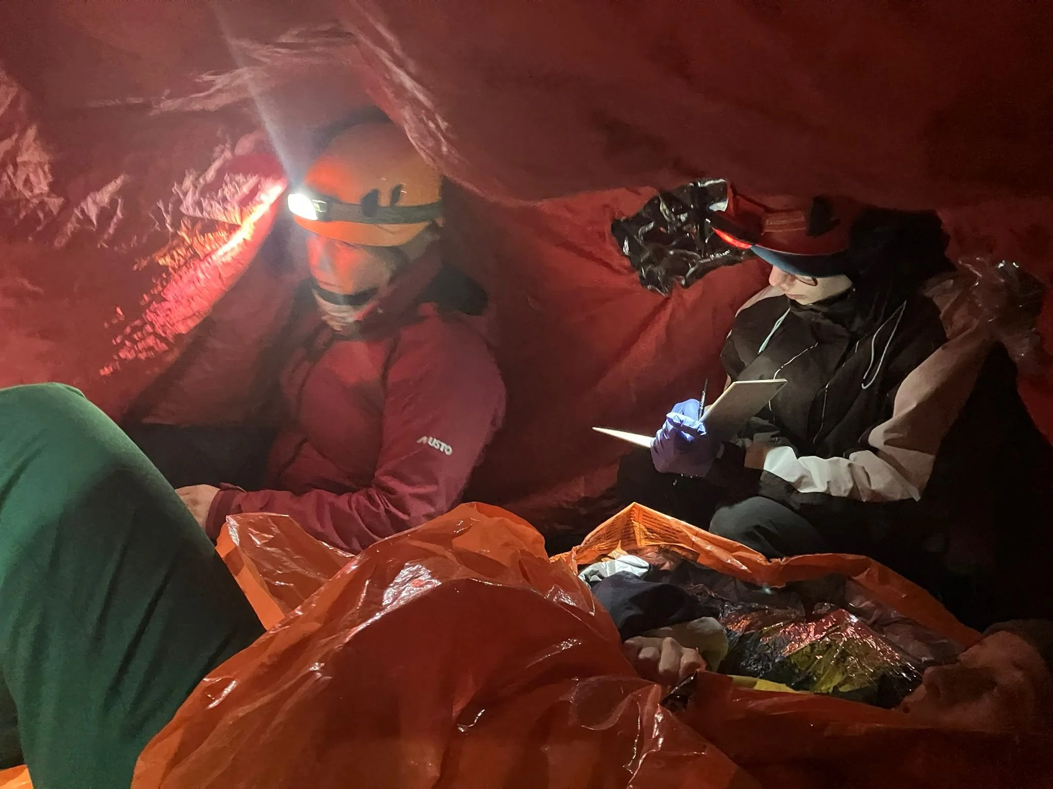 Two people inside a red cave or tent, wearing headlamps. One is taking notes on a small notepad and the other is resting or sleeping wrapped in a blanket.