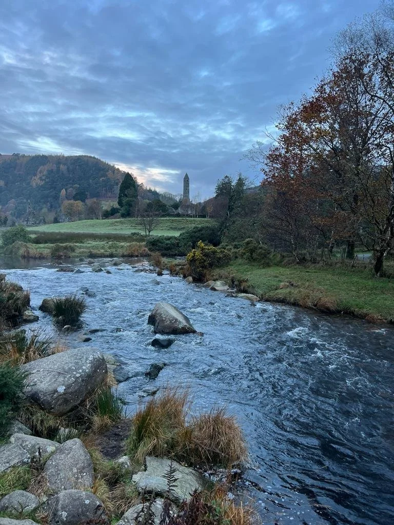 A flowing river with rocks along the banks, trees with autumn foliage, and a tall clock tower on a hill in the distance under a partly cloudy sky.