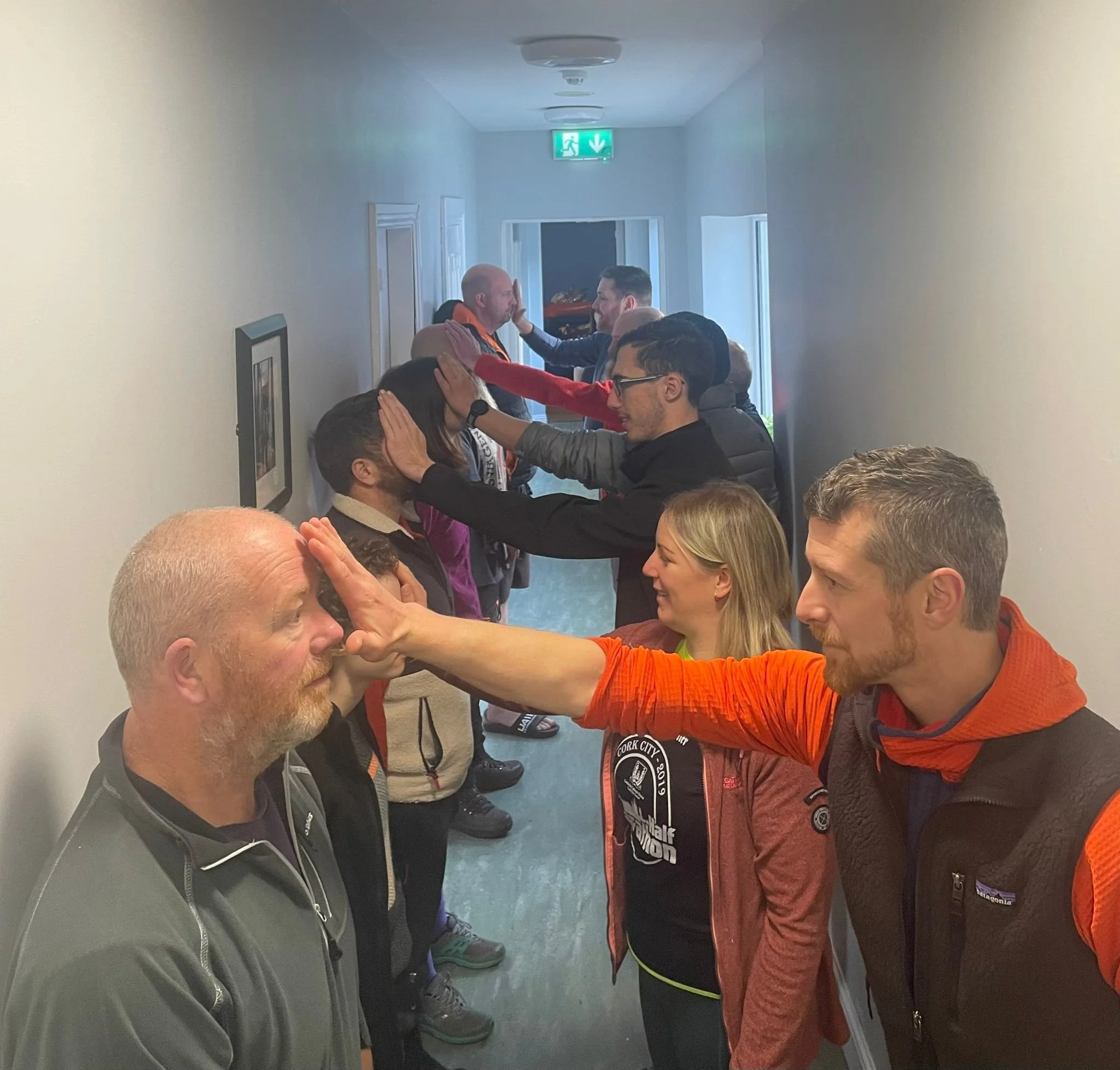 Group of people standing in a line in a hallway, participating in a meditative or relaxation activity, touching foreheads and faces of the person in front of them.