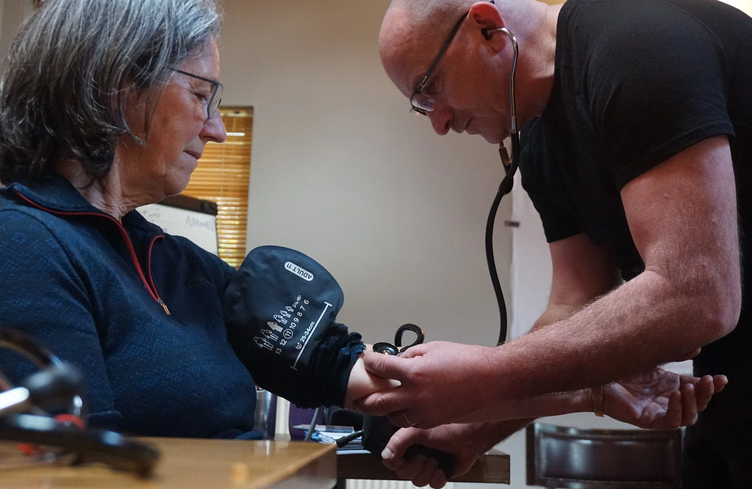 A healthcare worker measuring a patient's blood pressure with a sphygmomanometer while a healthcare professional monitors.