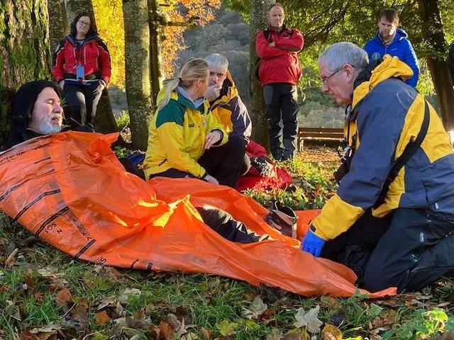 Emergency responders attending to a person lying on the ground outdoors, surrounded by trees and fallen leaves, during daytime.