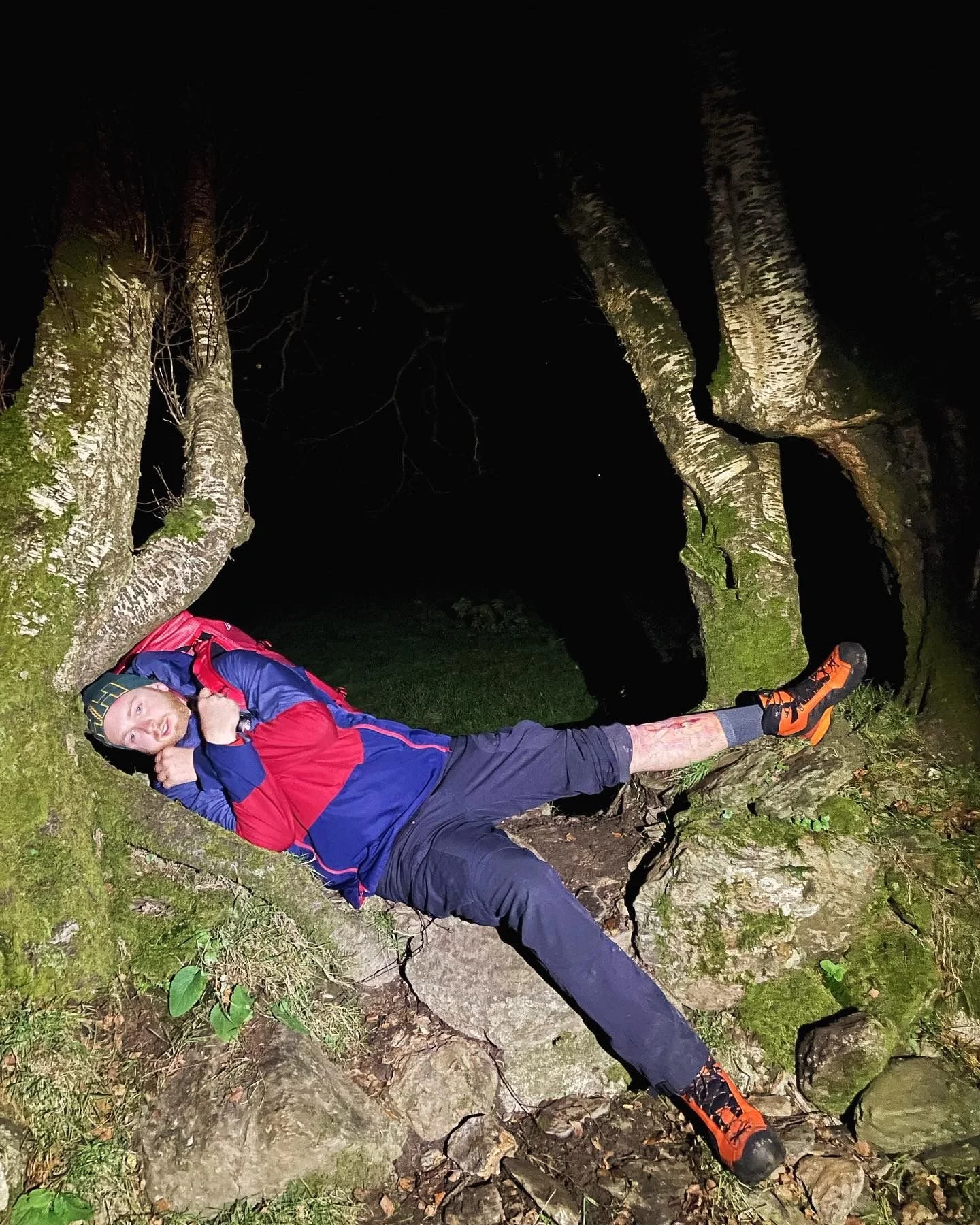 Person lying on a mossy, cracked tree trunk at night, with a backpack, wearing hiking gear and orange shoes.