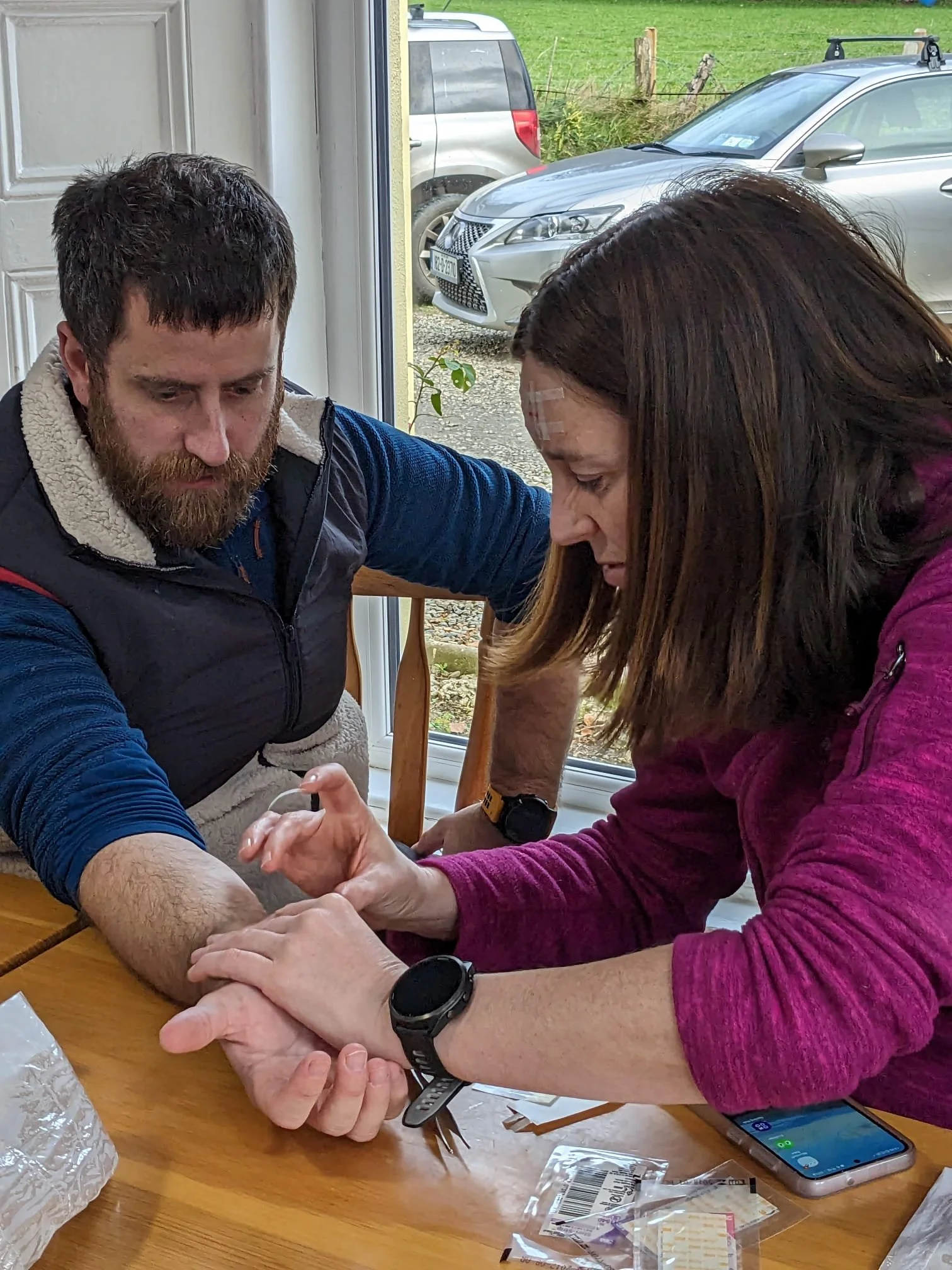 A woman is administering a medical test or injection to a man's arm while another person observes inside a home near a window, with cars parked outside.