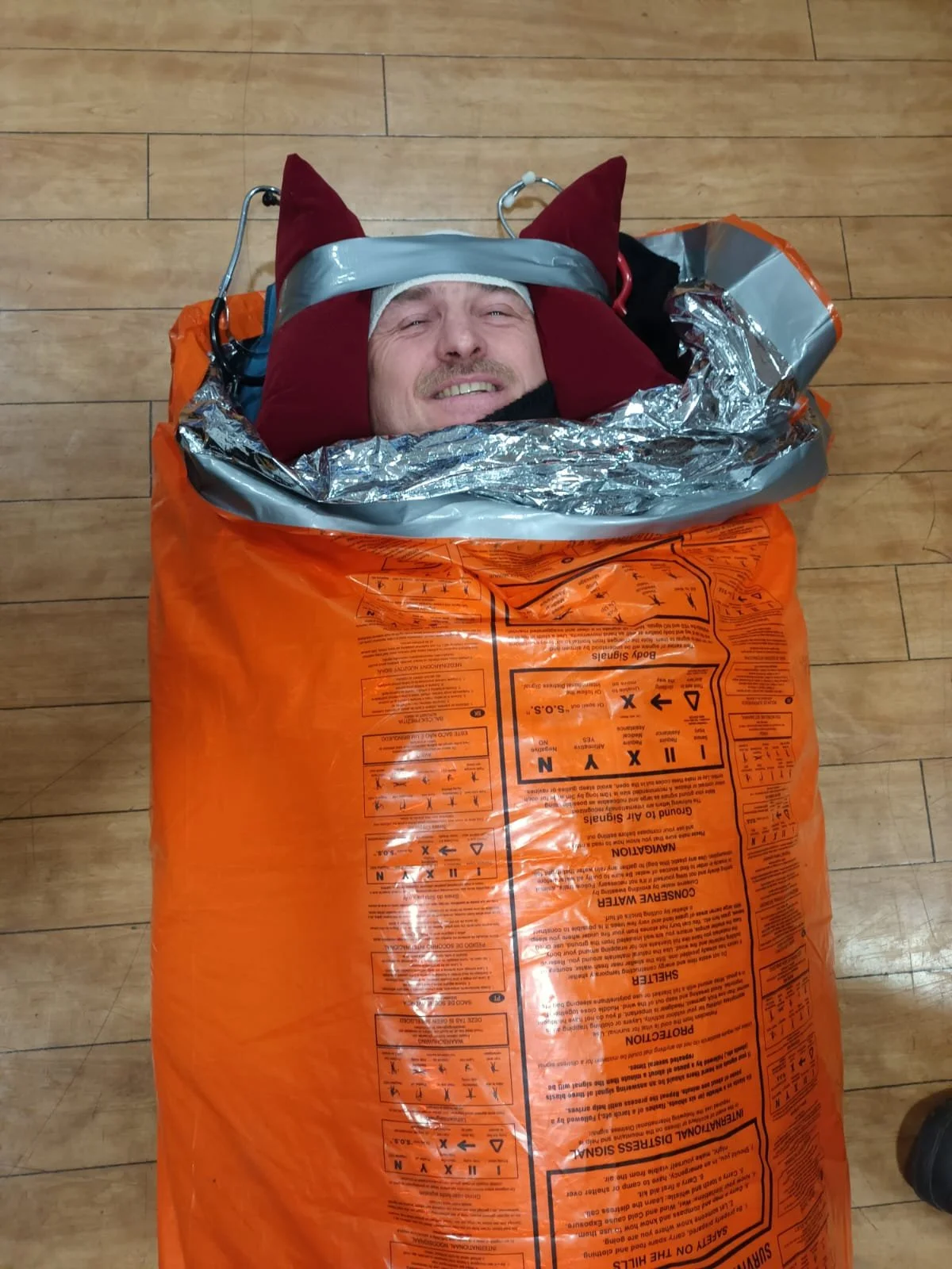 A man is lying inside a yellow emergency blanket bag with a smiling face, wearing a red hood with pointy parts and gray tape around his head, on a wooden floor.