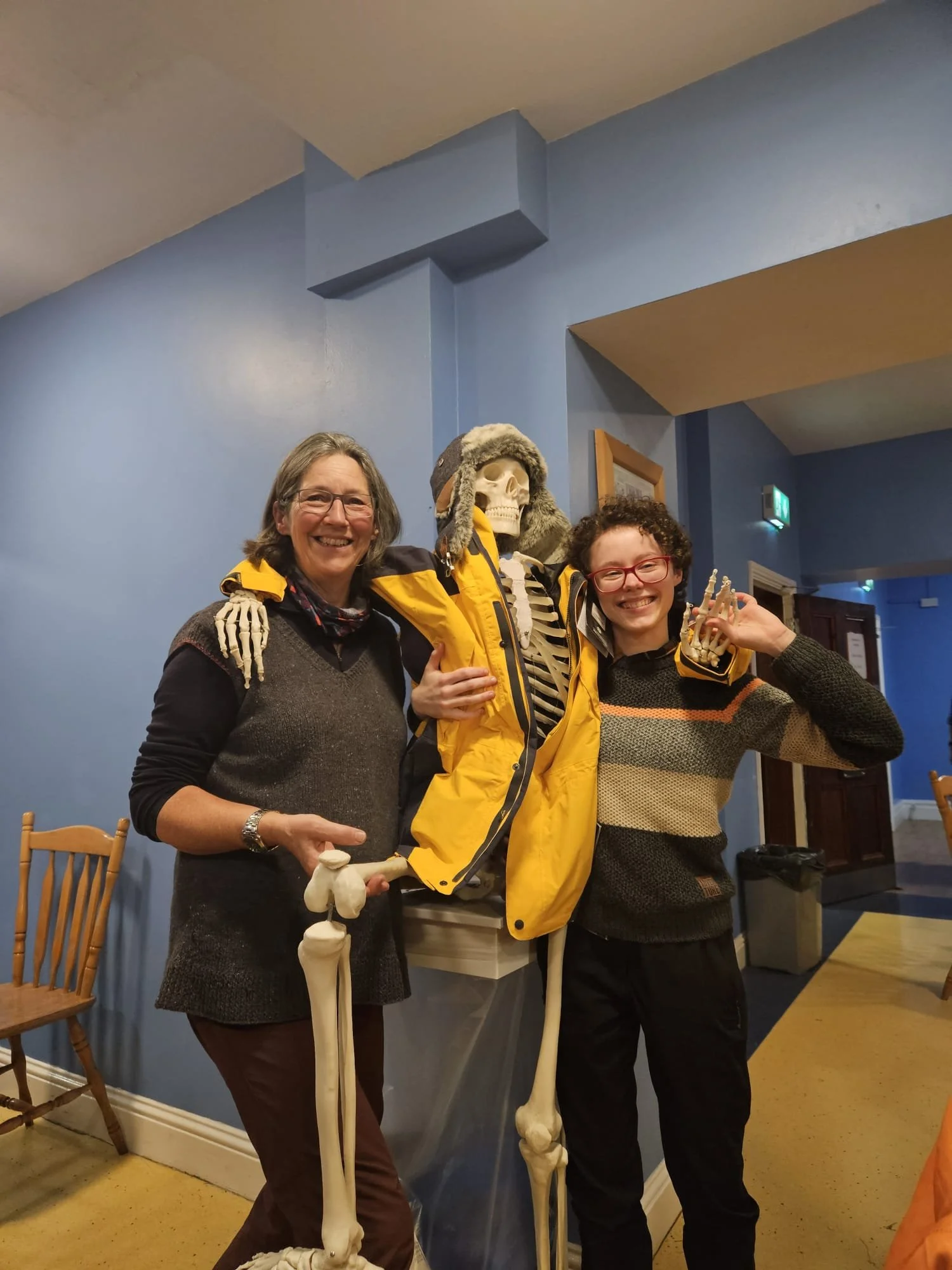 Two smiling women standing on either side of a life-sized skeleton in a yellow rain jacket, posing indoors against a blue wall.
