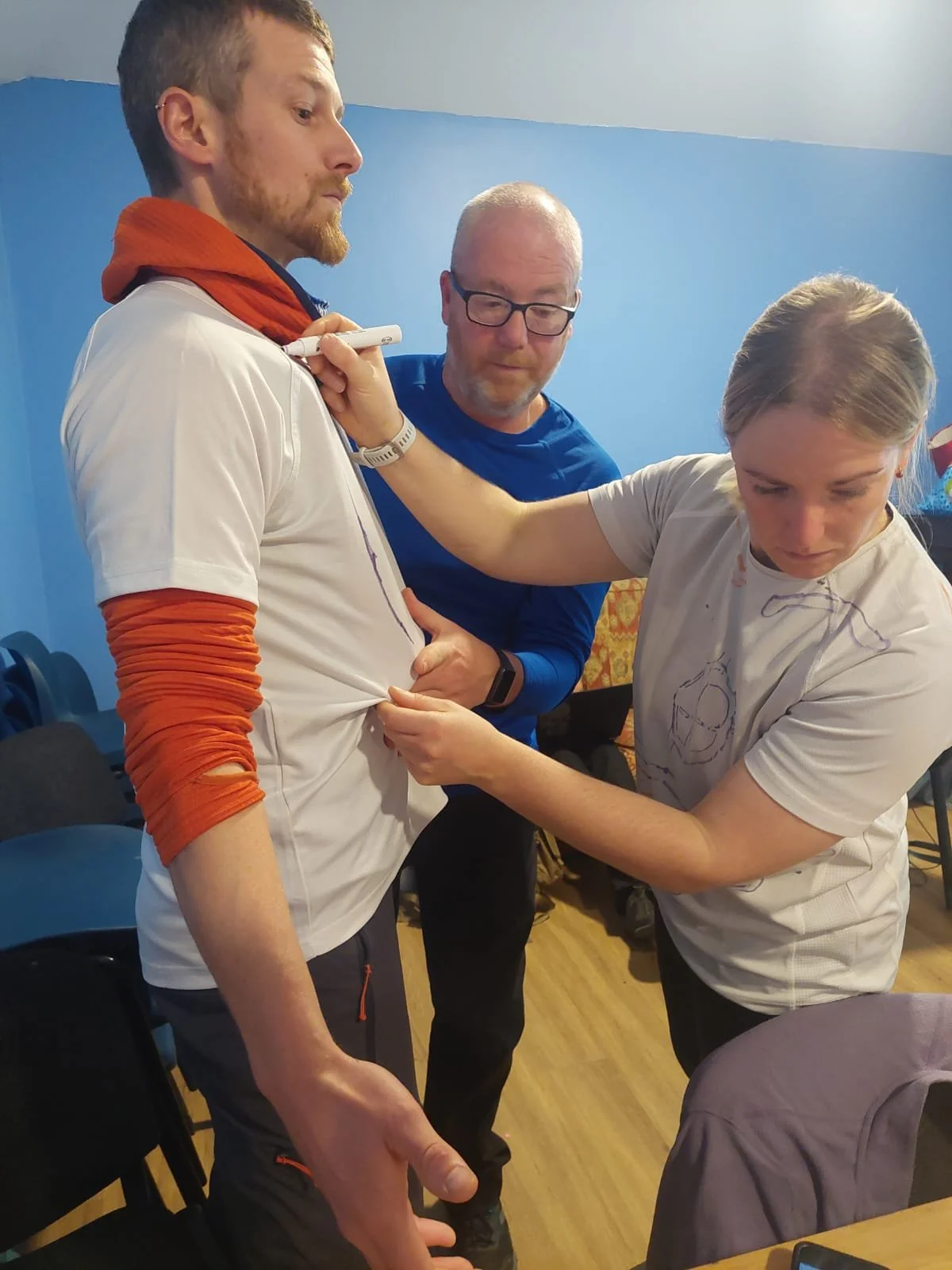 A group of three people in a room with a blue wall. One man is standing with a serious expression, wearing a white shirt with orange sleeves, and appears to be getting medical attention. A woman is examining his arm, while a man with glasses observes