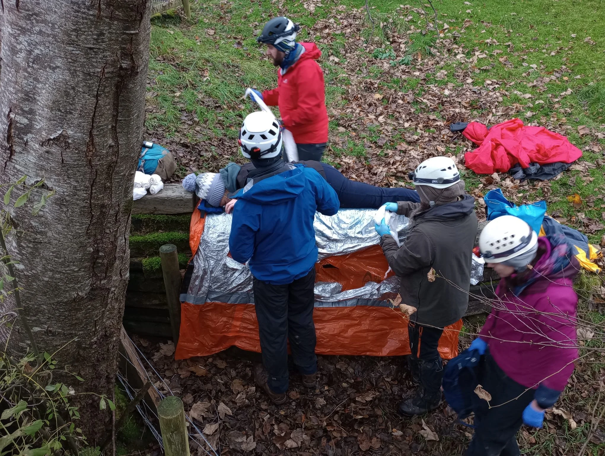 Group of people wearing helmets and outdoor gear attending to a person lying on a stretcher in a wooded area with fallen leaves.