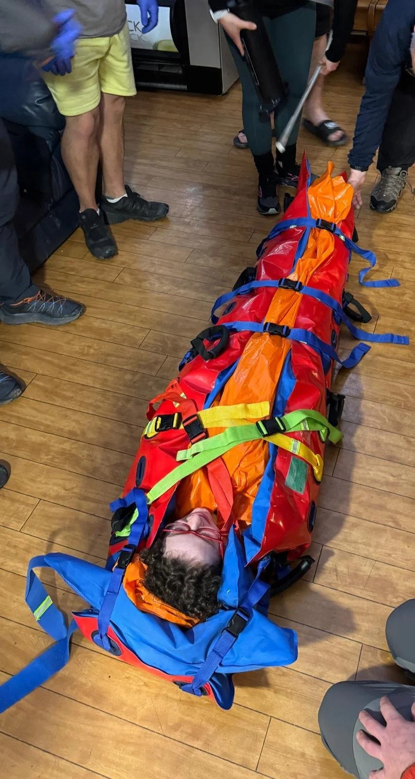 A person lying inside an emergency rescue stretcher with colorful straps, surrounded by several people, some of whom are wearing gloves, in a room with wooden flooring.
