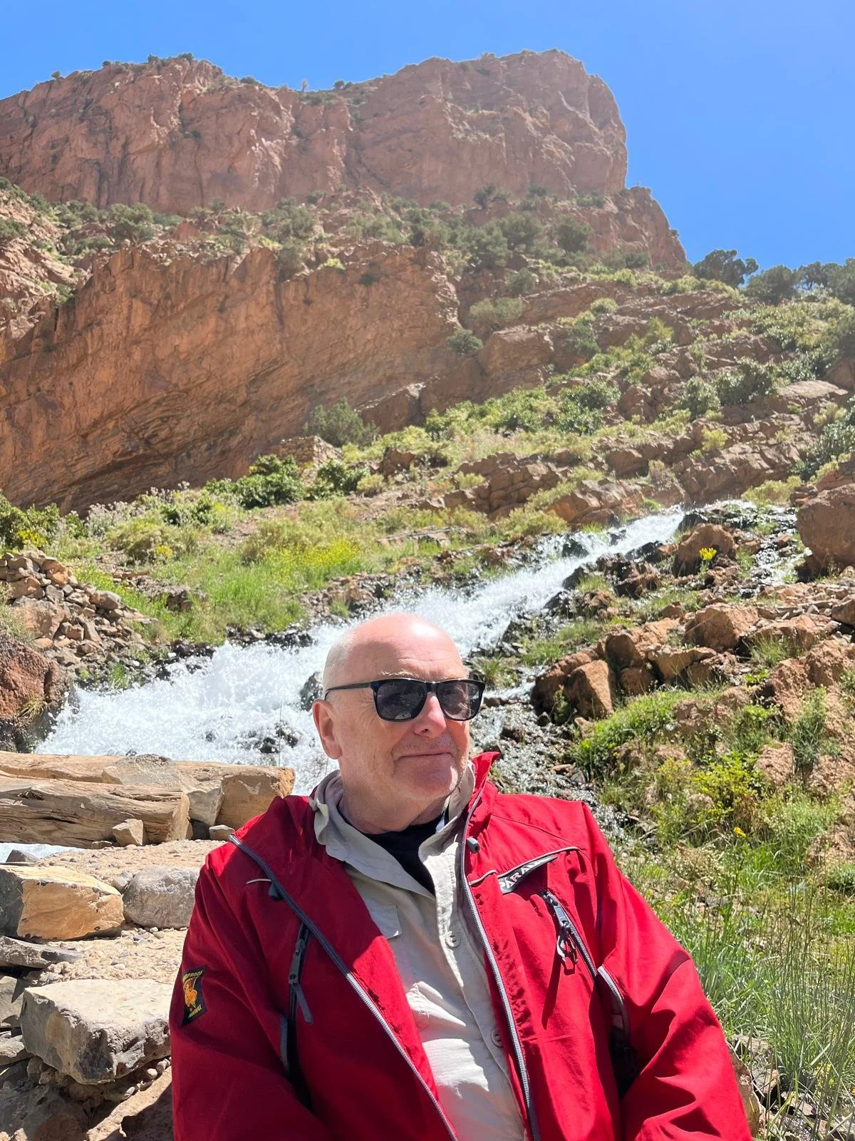 A man in a red jacket and sunglasses sitting on rocks near a flowing creek in a mountainous area with rocky cliffs and green vegetation under a blue sky.