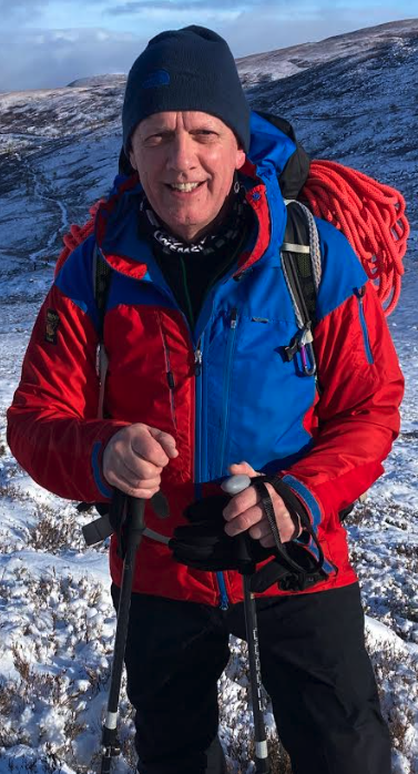 Man in winter gear hiking in snowy mountains, carrying a red rope on his shoulder.