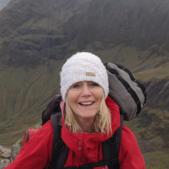 Woman with blonde hair wearing a gray knit beanie, red outdoor jacket, and carrying a backpack, smiling outdoors in a mountainous area.