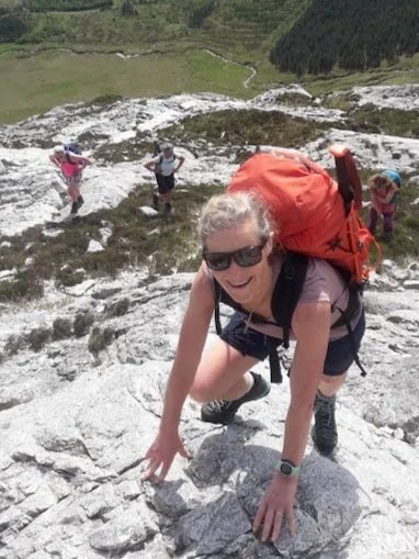 A woman climbing a rocky mountain with a large red backpack, accompanied by three children in the background, on a bright day with green hills and a small stream.
