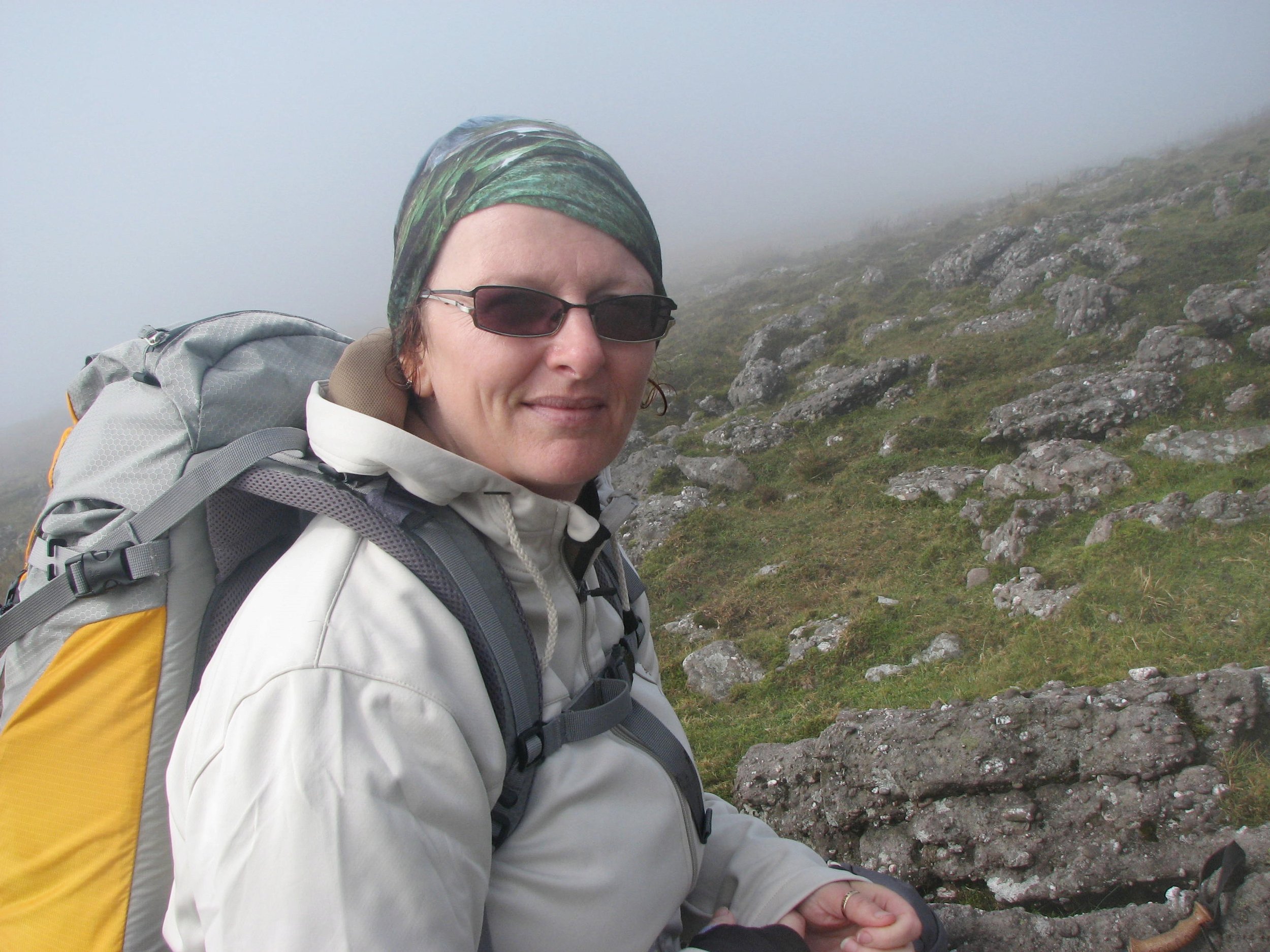 A woman wearing sunglasses, a green headscarf, a white jacket, and a yellow backpack, outdoors on a foggy, rocky hillside.