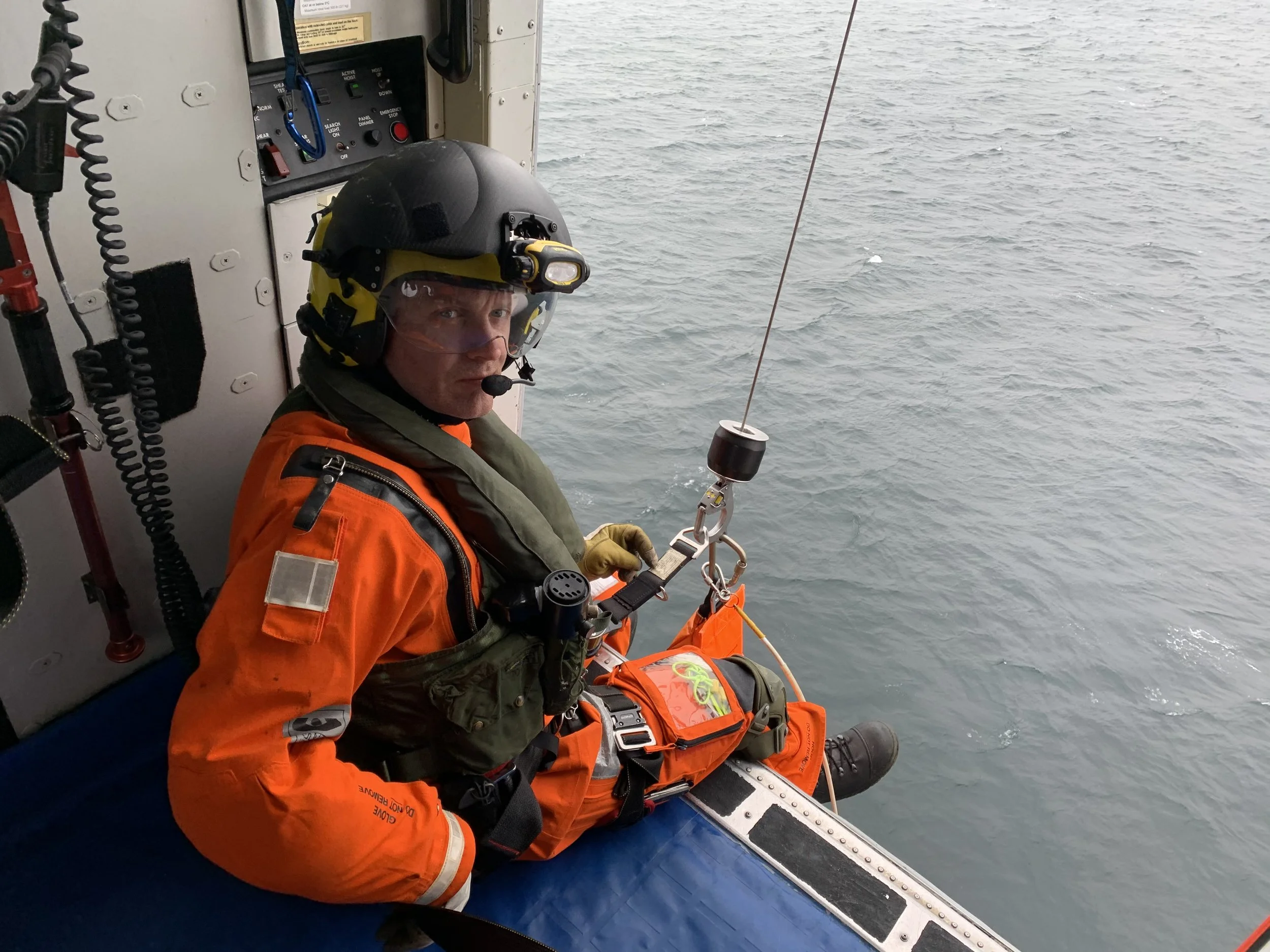 A man in an orange and green rescue uniform with a helmet and headset, seated at the edge of a vessel, looking at the camera, with the ocean in the background.