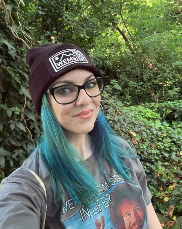 A young woman with blue-green hair wearing glasses, a maroon beanie with a WEMSI patch, and a t-shirt with a Redd Foxx image, standing in a lush green outdoor setting.