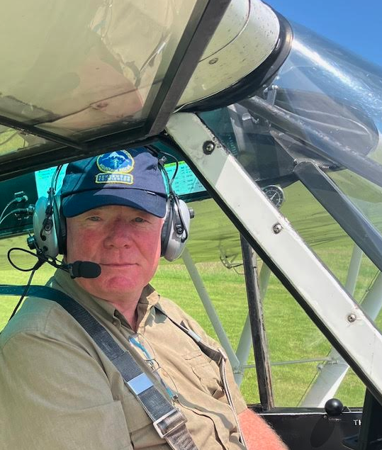 A man in pilot uniform wearing a blue cap and headset sitting in the cockpit of a small aircraft on a grassy runway.