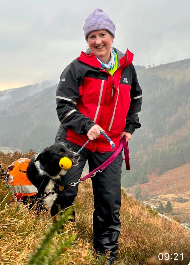 Woman hiking outdoors with a black and white dog, both dressed in hiking gear. The woman is smiling and wearing a purple beanie and a red and black jacket. The dog has a yellow ball in its mouth and is wearing a harness and an orange backpack.