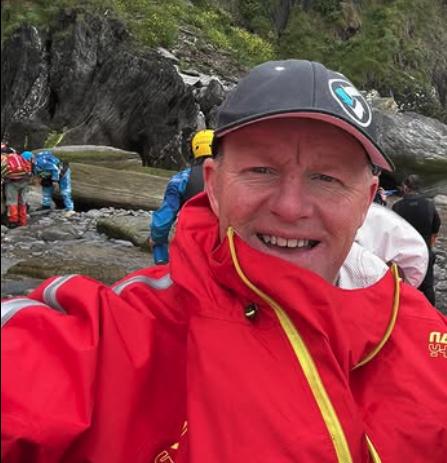 A man taking a selfie outdoors near a river, with several people in the background, some wearing life jackets and helmets, surrounded by rocks and greenery.