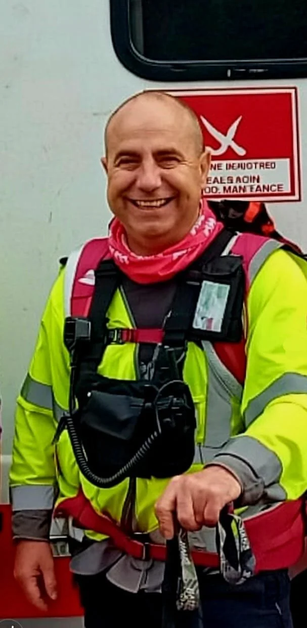 Smiling man in high-visibility yellow jacket with rescue gear, standing in front of a vehicle with a red warning sign.