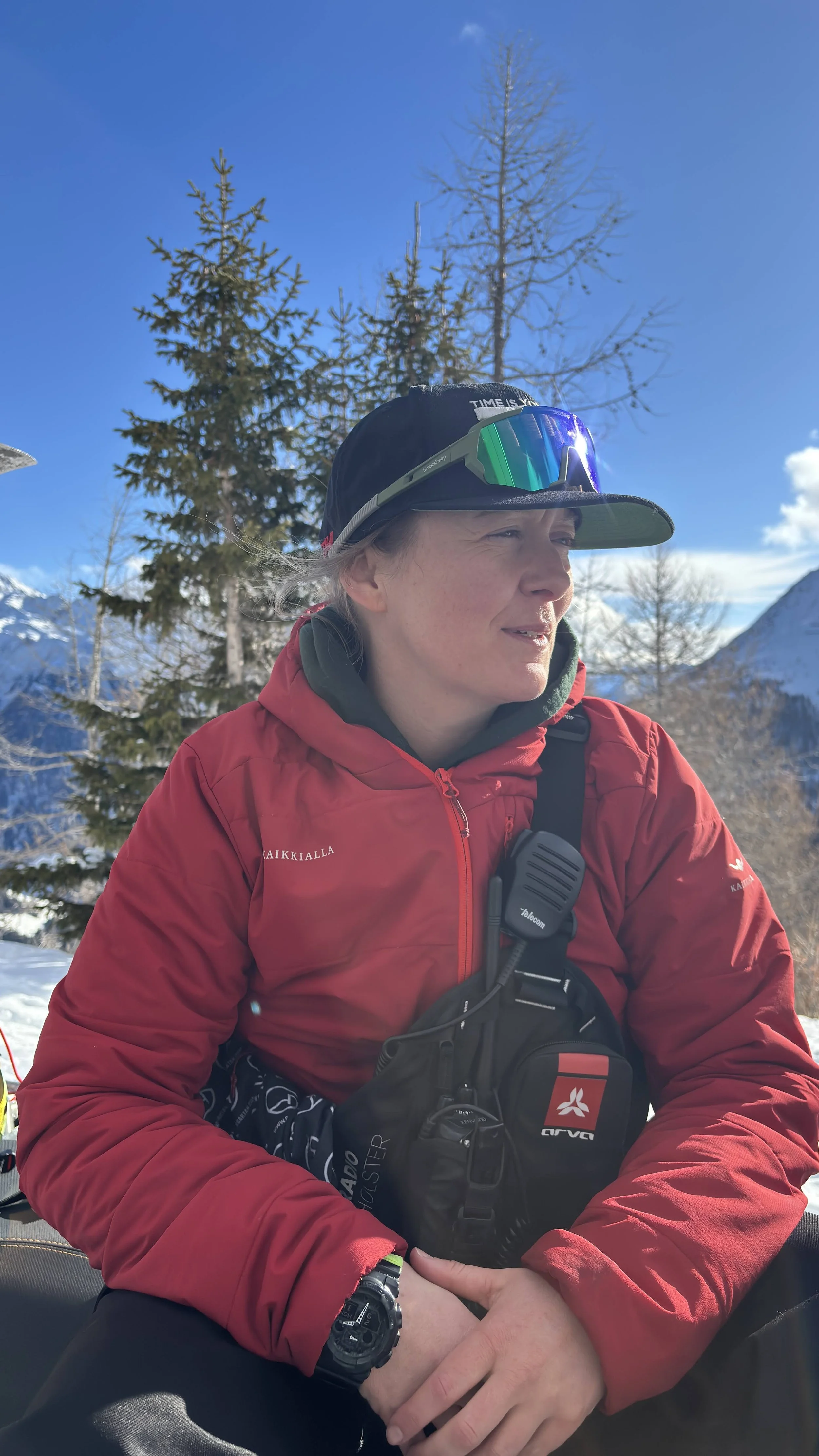 A woman dressed in ski gear standing on snow with a ski pole upright in her hand, mountainous snowy landscape in the background, cloudy sky, and the sun partially obscured.