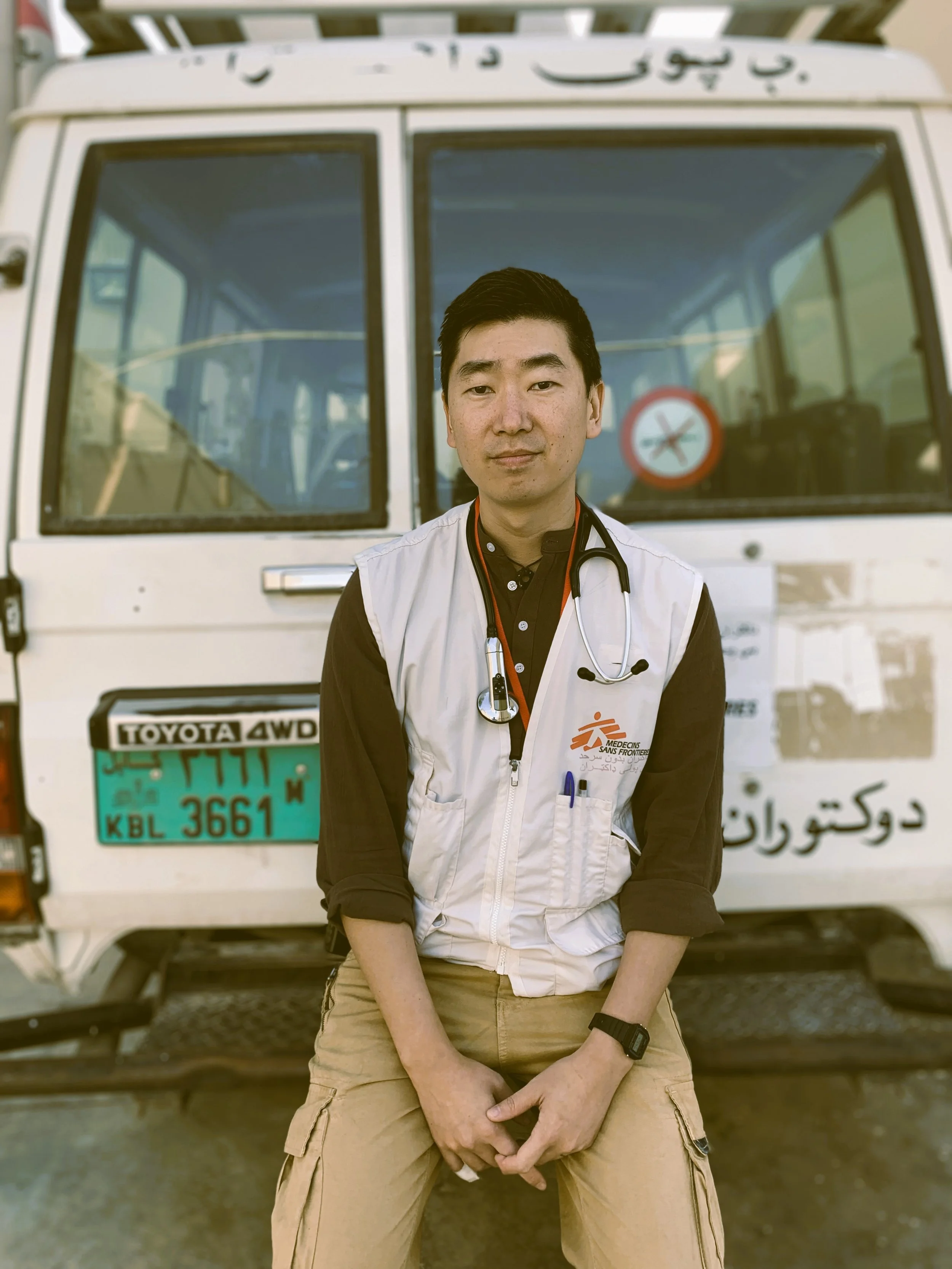 A young male healthcare worker with a stethoscope around his neck sits in front of a vehicle with a sign and writing in Arabic. He is wearing a black shirt with the sleeves rolled up, a white vest with the logo of Médecins Sans Frontières, and a black watch. The vehicle behind him has a license plate with English and Swahili characters and a 'no smoking' sign on the windshield.