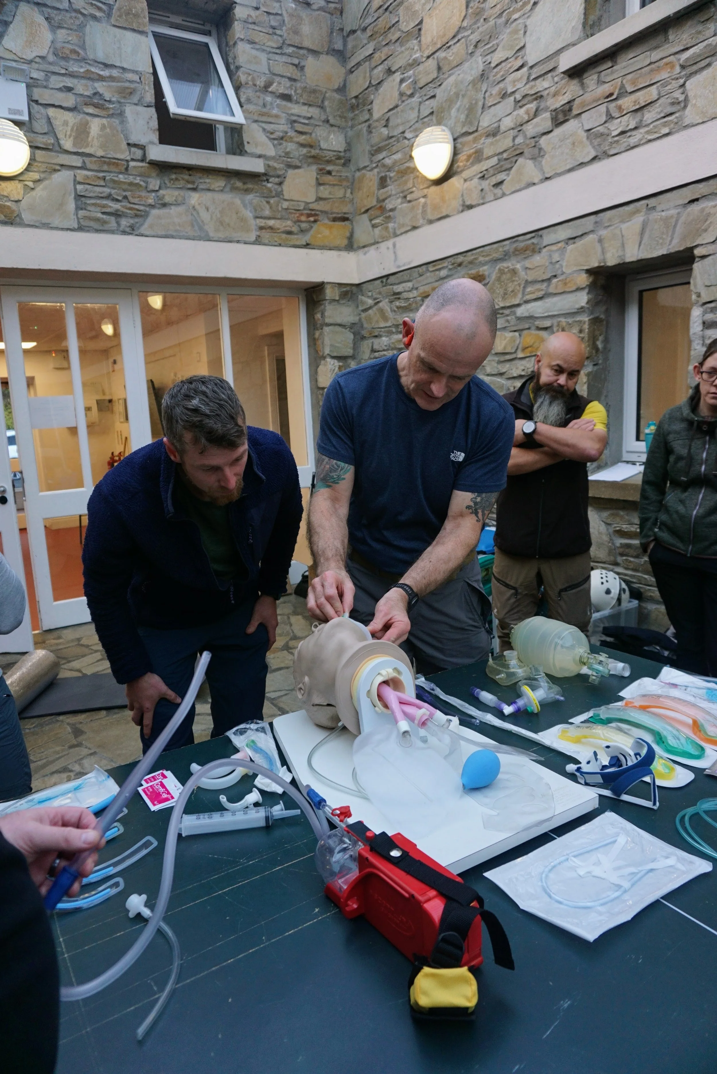 A group of people working on a medical training simulation with a human mannequin head, medical tools, tubing, and supplies on a table inside a stone-walled room.