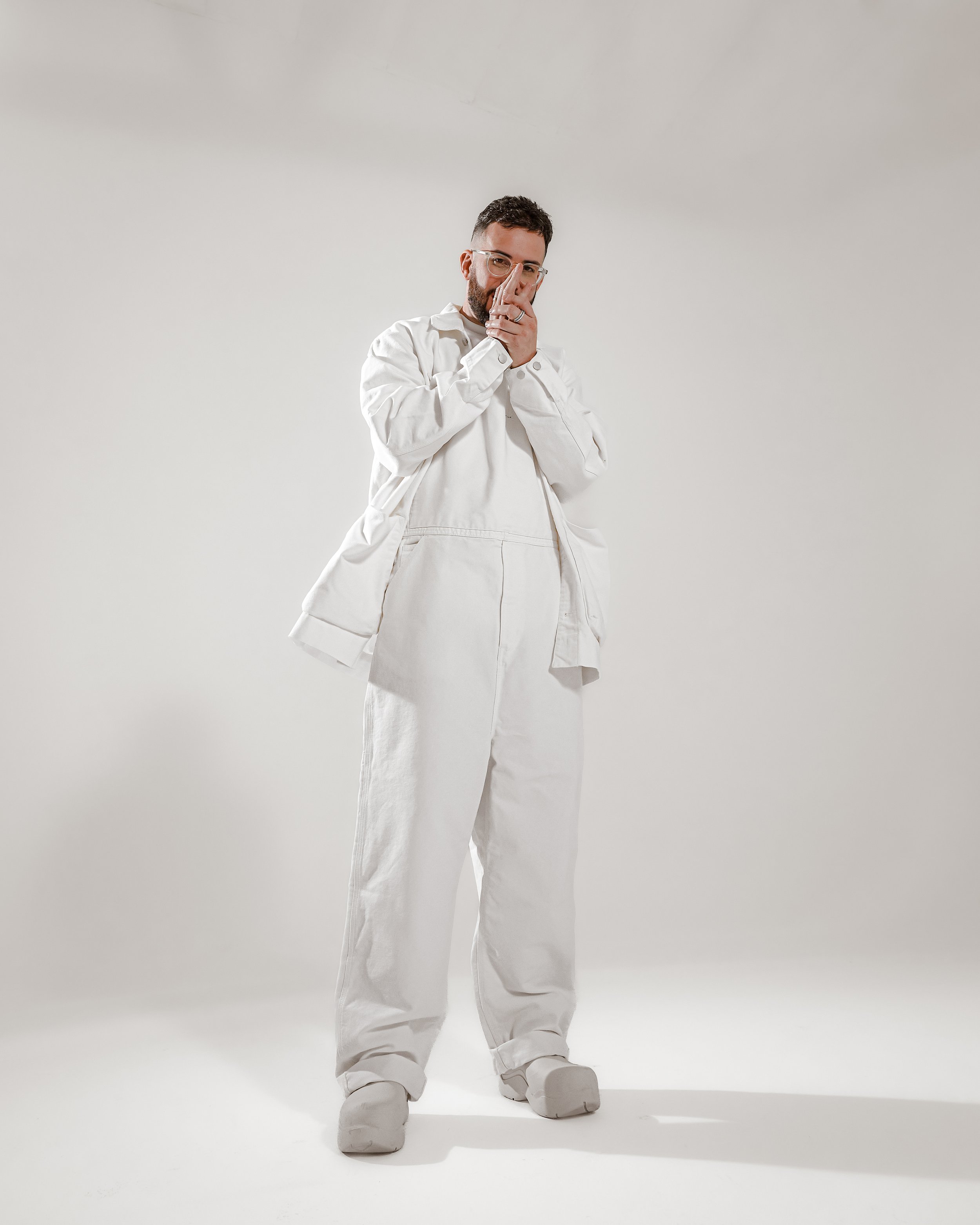 A man in white oversized clothing, including baggy pants, a large shirt, and sneakers, stands against a plain light gray background, with his hands near his face, wearing glasses and displaying a thoughtful expression.