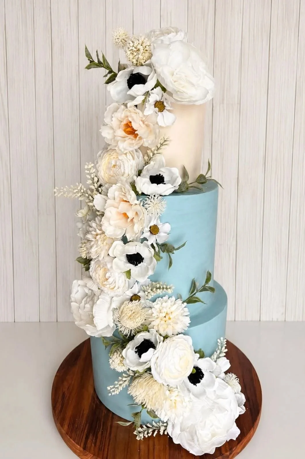 A three-tiered cake with a bottom blue layer, a middle white layer, and a top beige layer, decorated with white, cream, and black flowers cascading down one side, on a round wooden cake stand.