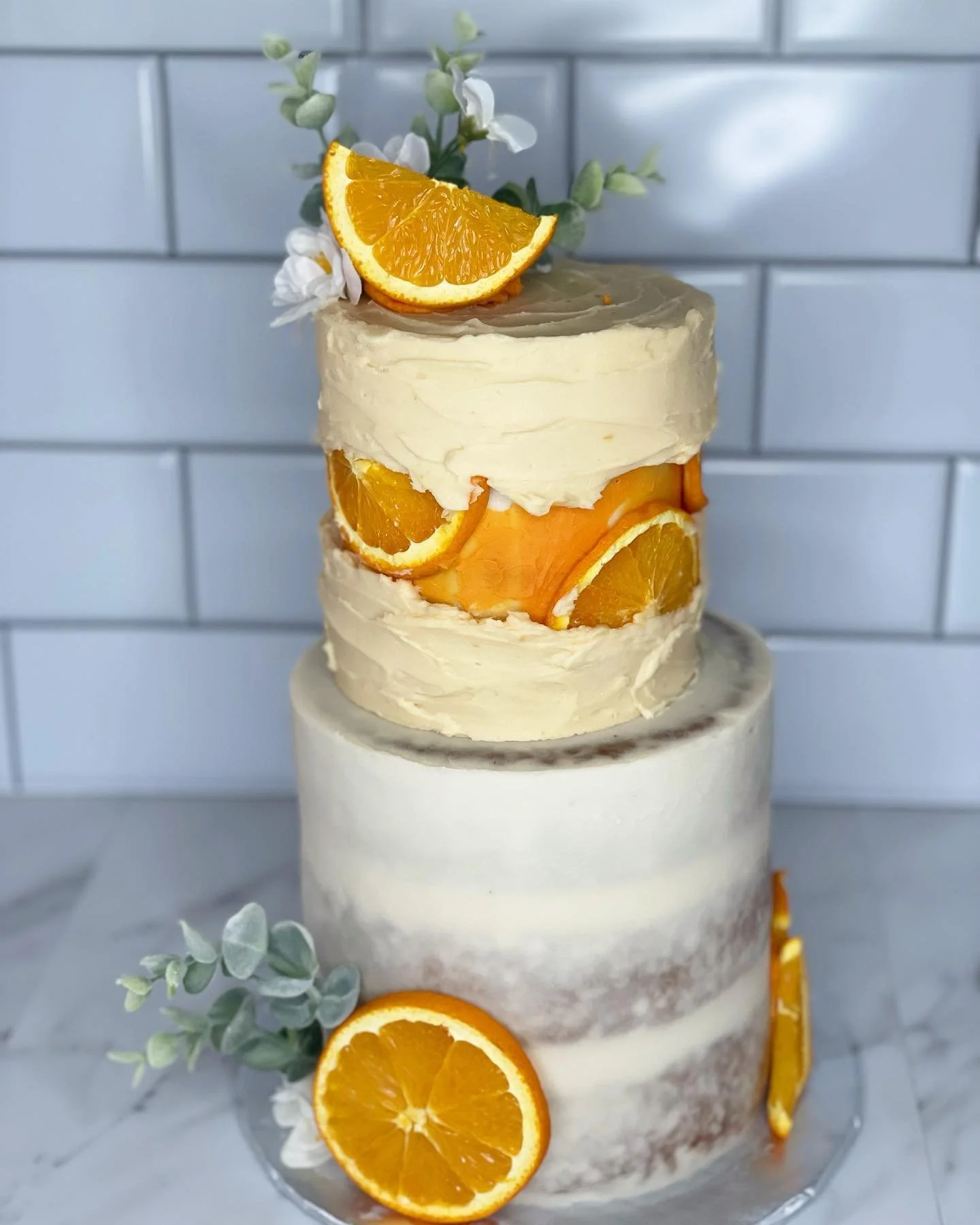 A three-tiered cake decorated with orange slices and white flowers, with a sprig of green foliage, set against a white brick background.
