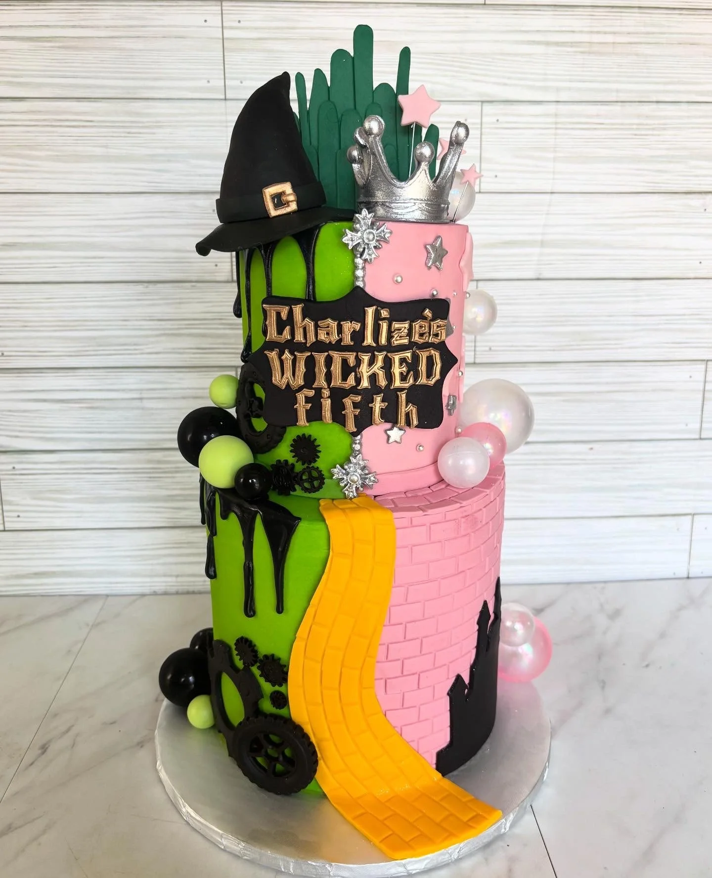 A colorful birthday cake with a dark witch hat, green and pink sections, a silver crown, and a yellow fondant path, decorated with stars, beads, and gear shapes, and the phrase 'Charlize's Wicked Fifth' on a black banner.