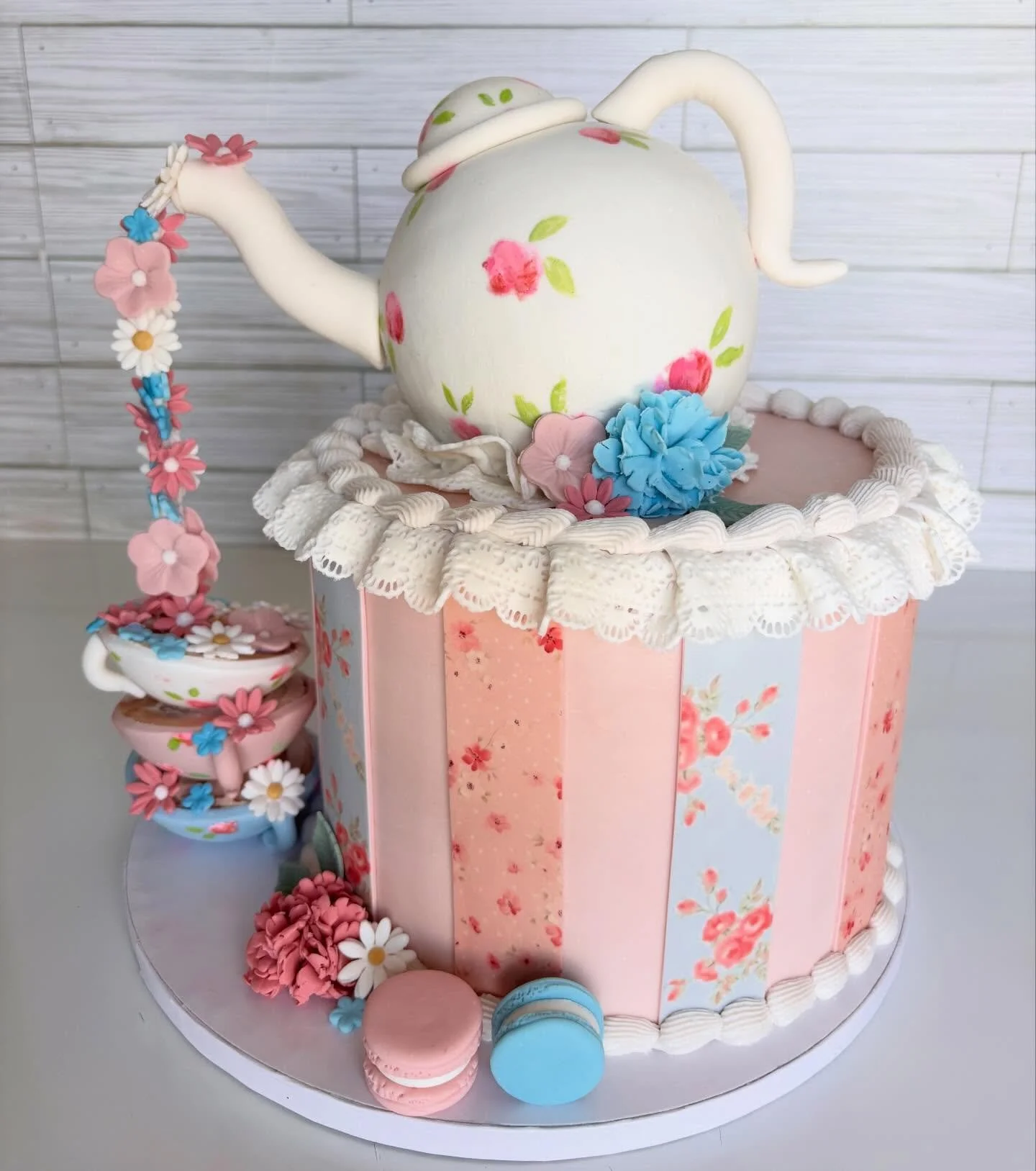 Decorative teapot with a floral design and a branch with flowers, placed on a pink and blue floral-printed cake stand along with teacups and macarons.