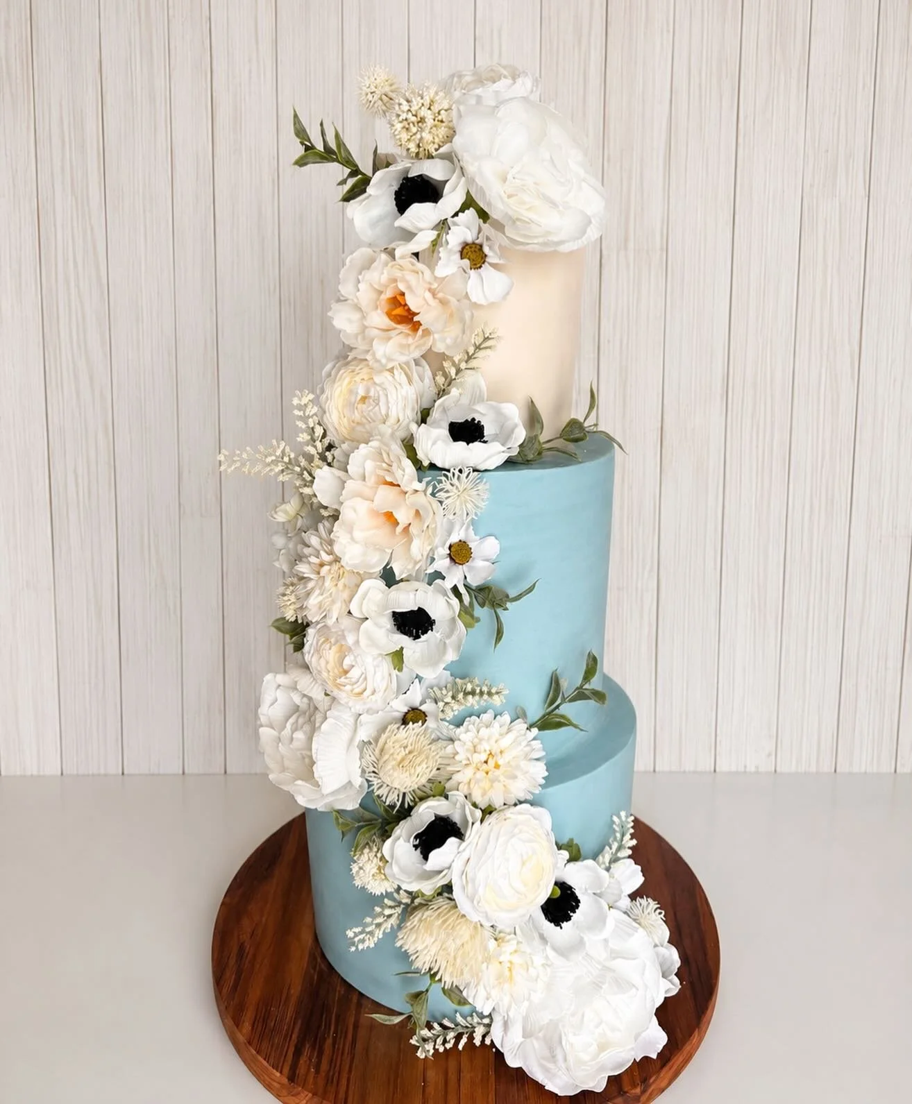 A three-tiered cake with light blue icing, decorated with cascading white and cream-colored flowers, on a wooden cake stand against a white wall with vertical paneling.