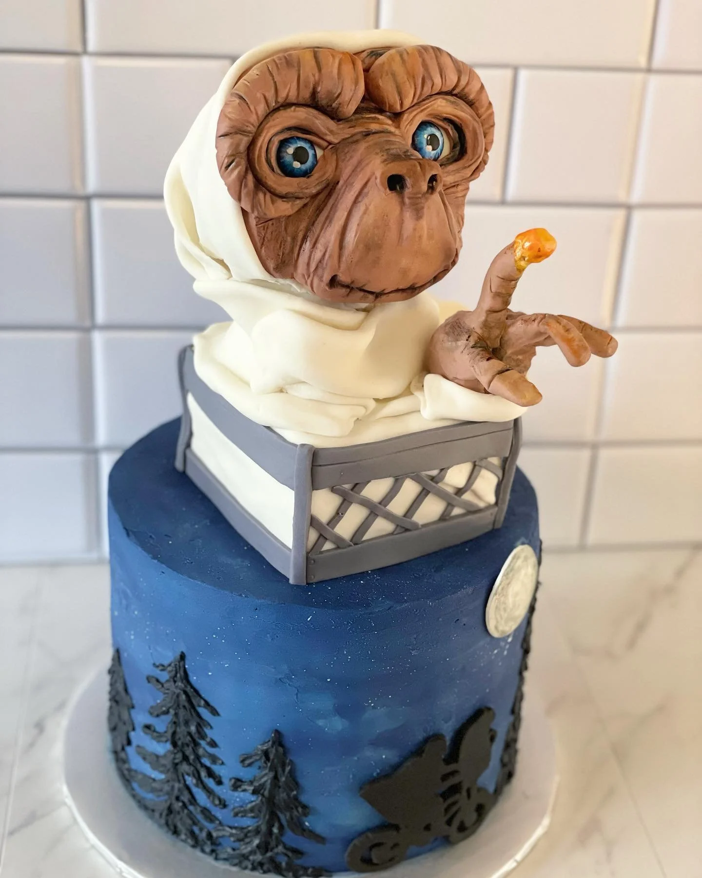 Cake featuring a detailed edible sculpture of a dog's face, wearing a white hoodie, with a small basket and urban cityscape design on the base, set against a tiled background.