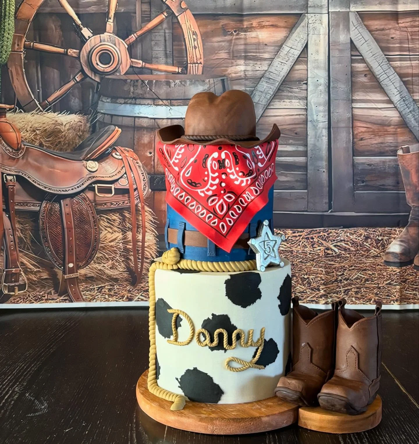 Cowboy-themed birthday setup with a cake that has a cow print pattern and the name 'Dany' on it, a pair of cowboy boots, a cowboy hat, a sheriff badge, and a red bandana, set against a rustic barn wall backdrop.