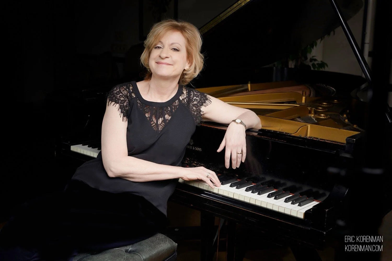 A woman with blonde hair in a black lace top sitting at a grand piano, smiling with one arm resting on the piano.