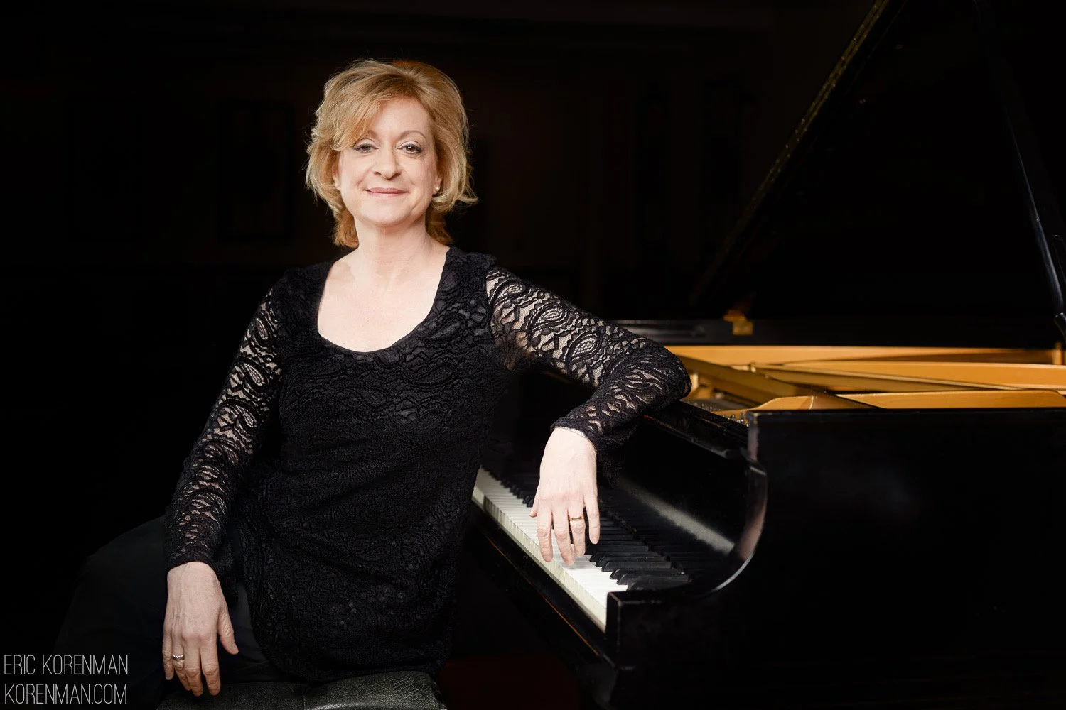 A woman with short red hair, wearing a black lace long-sleeve top, sitting by a grand piano in a dark room.