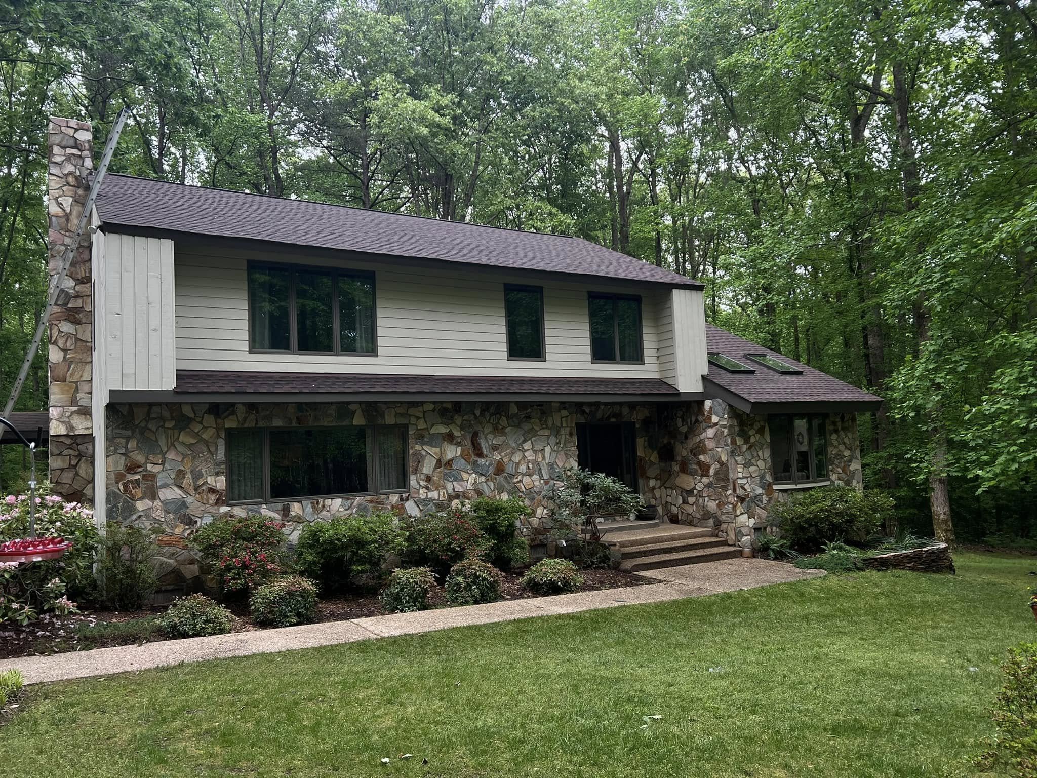A two-story house with stone and white siding exterior, multiple windows, surrounded by greenery, trees, a garden with bushes and flowers, and a paved pathway leading to the entrance.