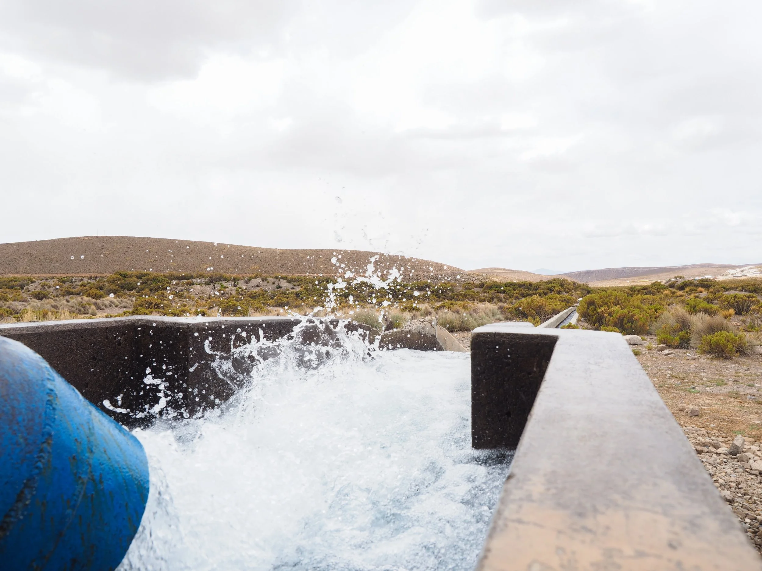 Agua saliendo de una tubería en un paisaje desértico con colinas y vegetación escasa bajo un cielo nublado.