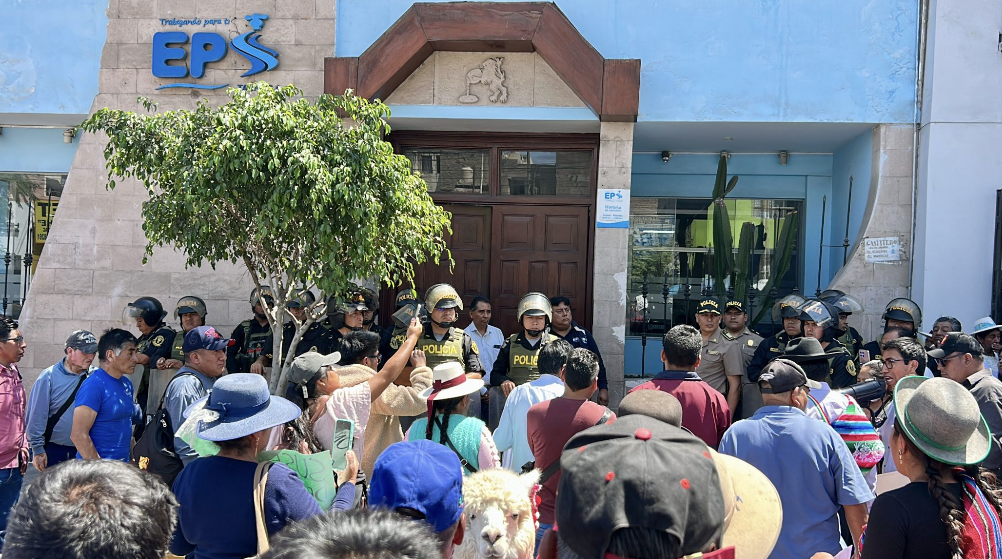Grupo de personas, incluyendo policías con cascos y civiles, congregados frente a un edificio azul con un letrero que dice 'Trabajando para ti EPS'.
