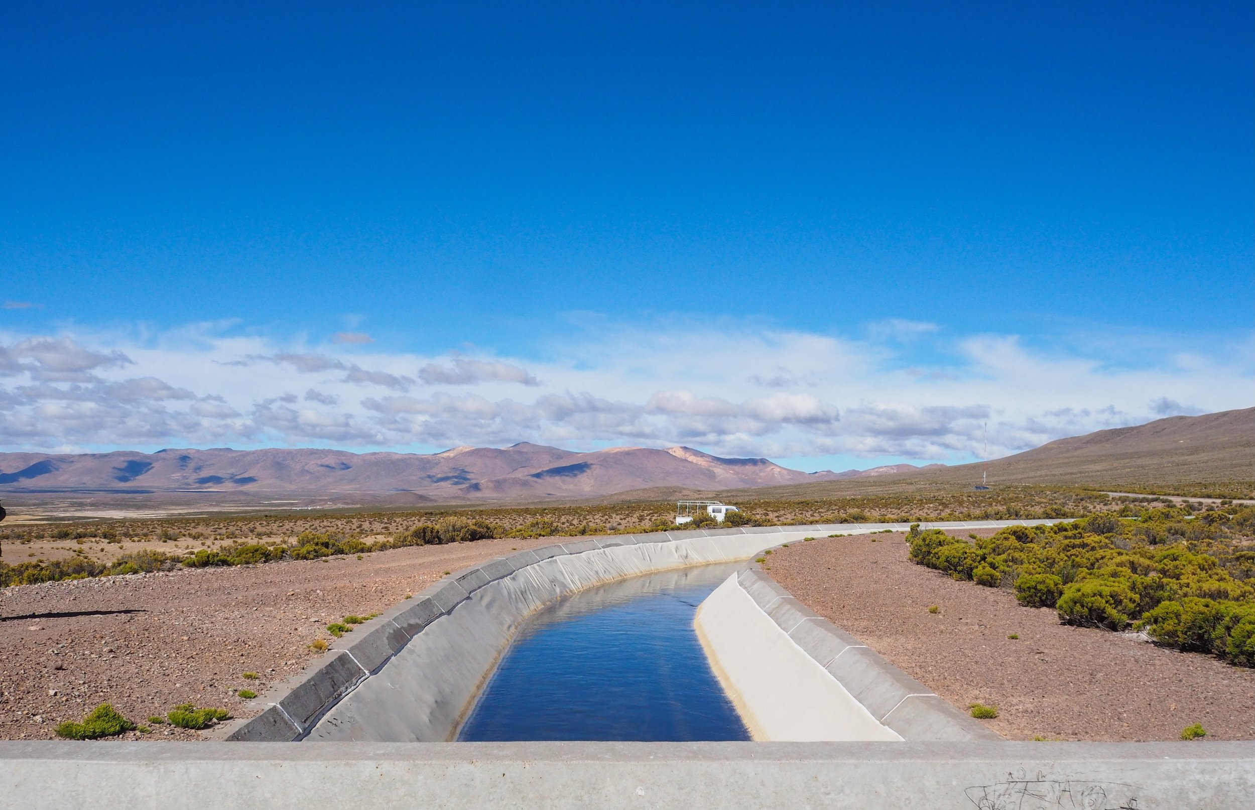 Vista de un paisaje árido con montañas al fondo, un canal de agua y vegetación dispersa en primer plano, cielo despejado con algunas nubes.
