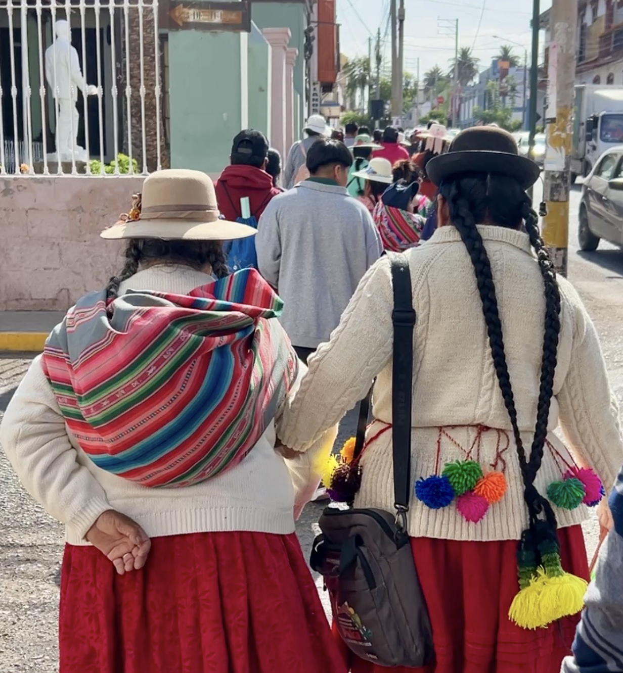 Personas vestidas con ropa tradicional, atravesando una calle en una ciudad, con edificios y vehículos en el fondo.