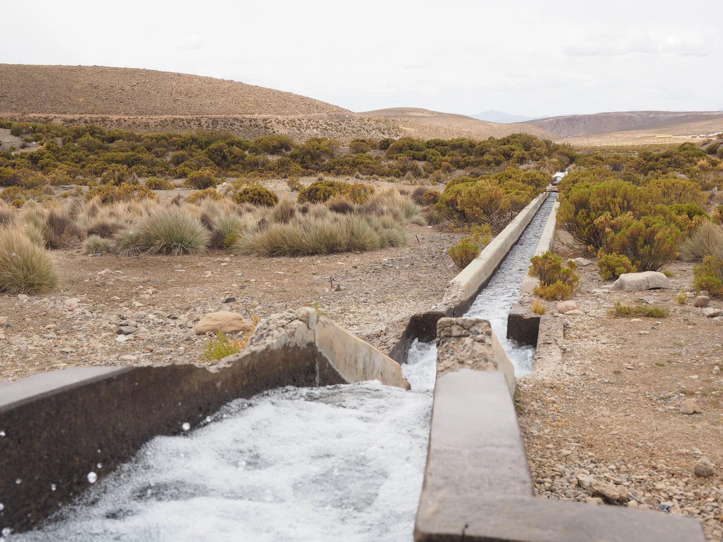 Vista de una tubería de agua que corre por el suelo, con colinas áridas y un cielo nublado en el fondo.